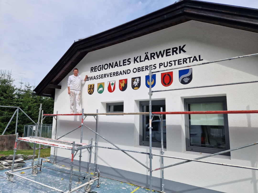 Handwerker auf einem Baugerüst vor dem hellen Gebäude des Regionalen Klärwerks Anras; über der Fassade die Aufschrift „Regionales Klärwerk – Abwasserverband Oberes Pustertal“ mit den Wappen der acht Mitgliedsgemeinden.