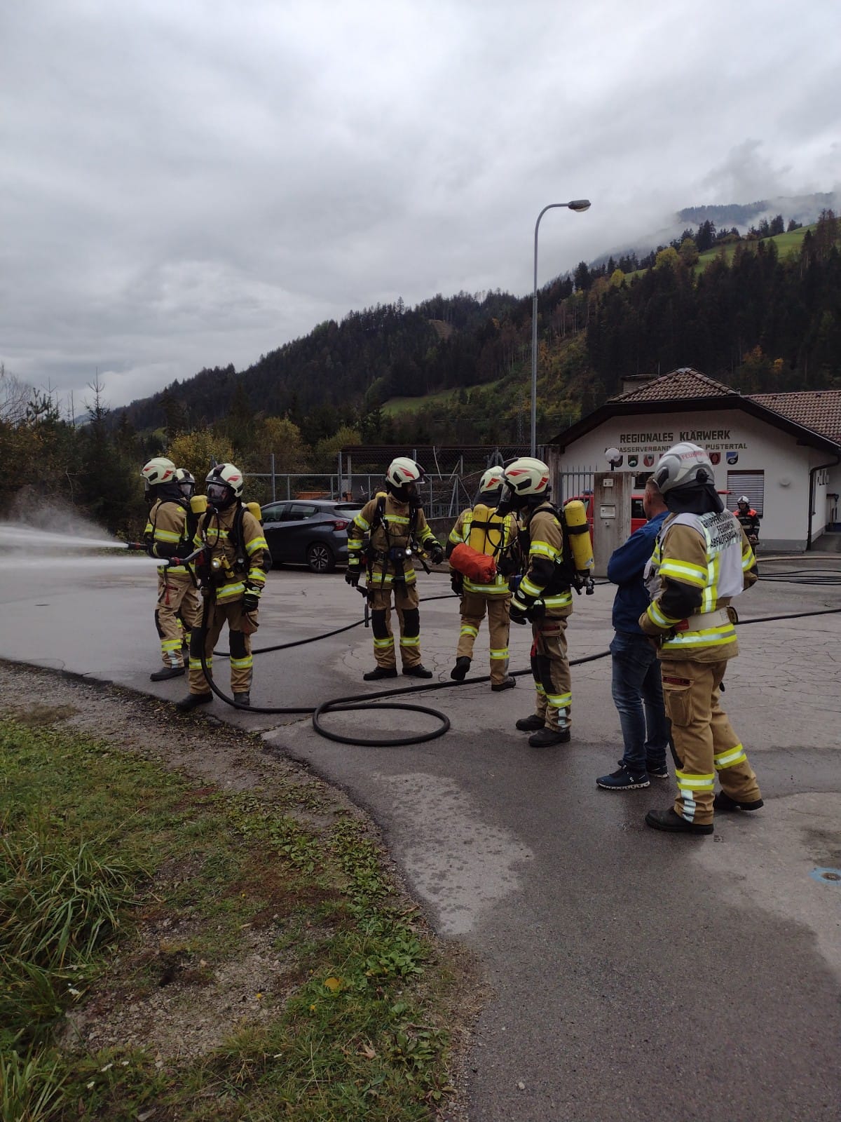 Mannschaft in Einsatzkleidung mit Atemschutzgeräten auf dem Vorplatz des Klärwerks, daneben ein Schlauch und eine Wasserwand aus einem Strahlrohr.