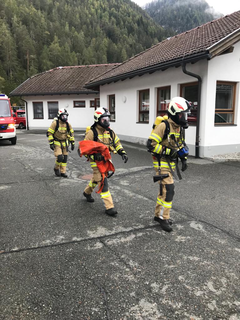 Drei Feuerwehrmänner mit Atemschutzmasken und Pressluftflaschen gehen mit Ausrüstung am Betriebsgebäude des Klärwerks vorbei.