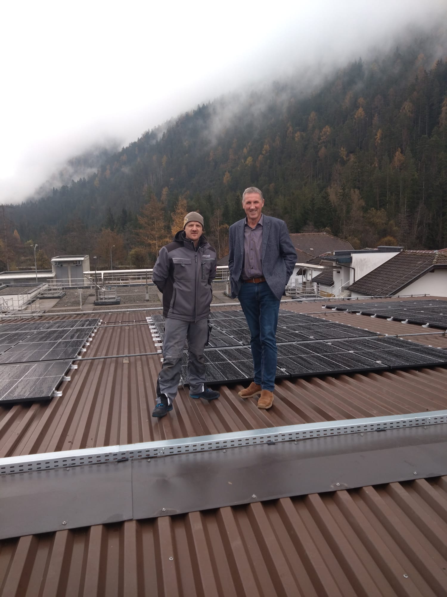 Zwei Männer stehen auf dem Dach der Kompostieranlage zwischen den flach aufgestellten Photovoltaikmodulen; im Hintergrund ein bewaldeter Hang in herbstlichem Nebel.